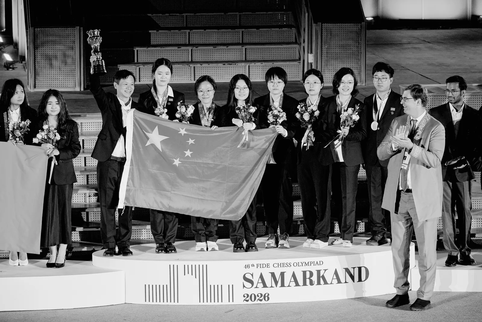 China's women's team on the Olympiad podium in Samarkand, holding the Chinese flag as a team official raises the trophy.