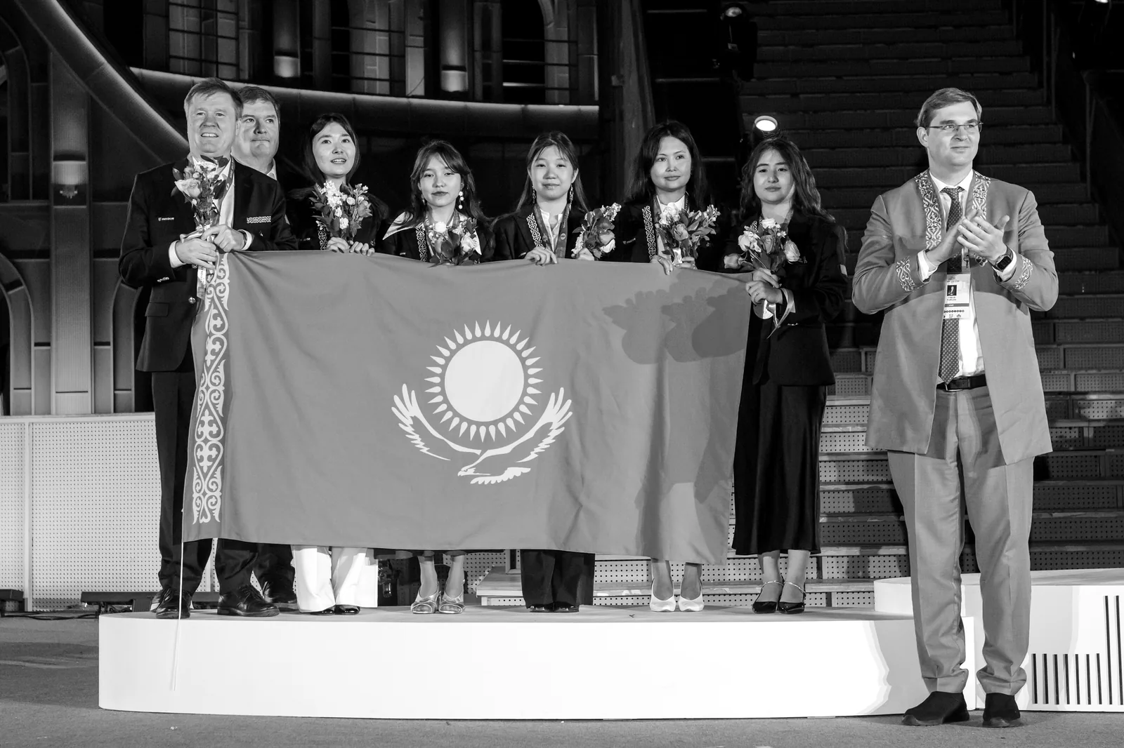 Kazakhstan's women's team on the podium in Samarkand, holding the national flag with their silver medals and flowers.