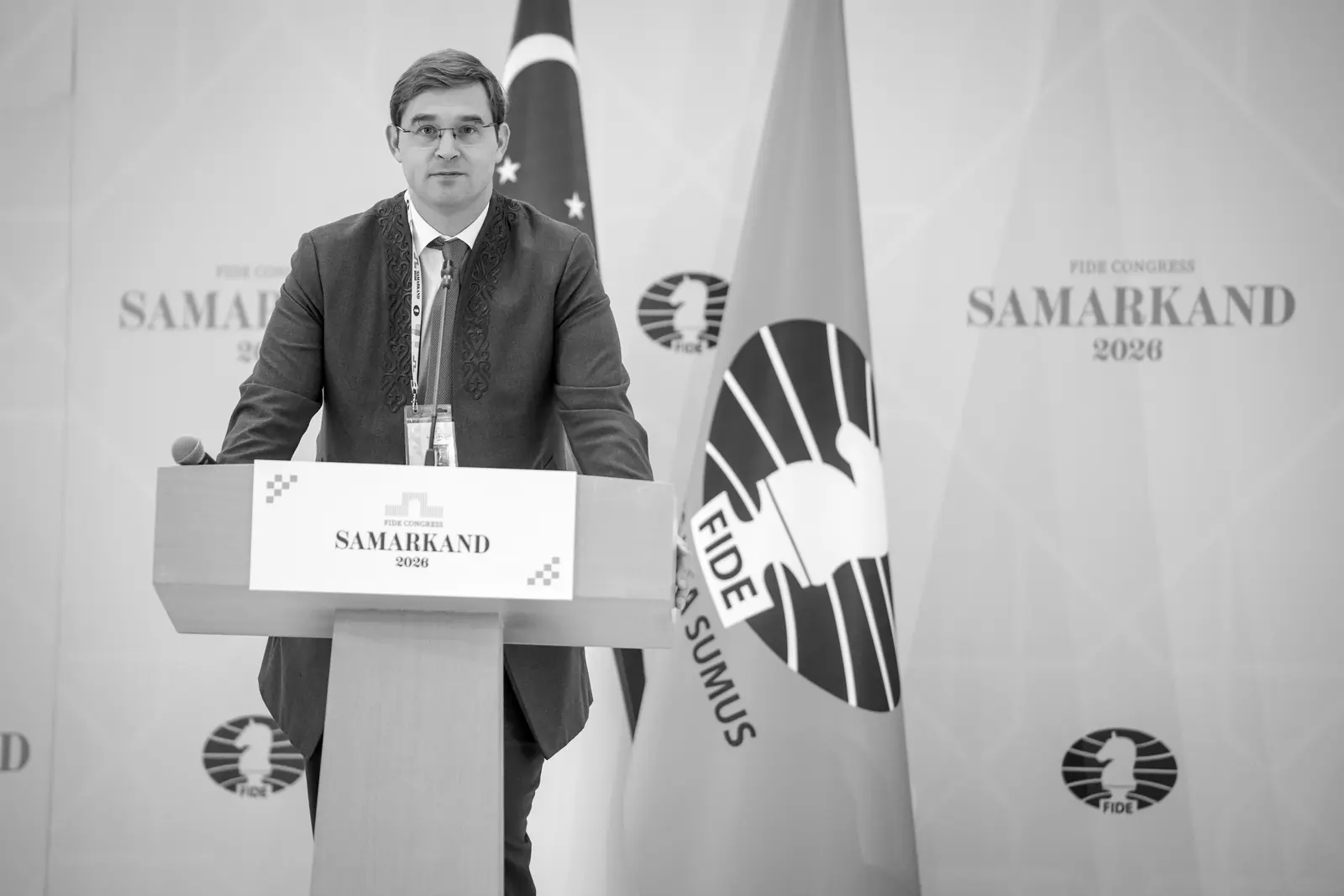 Timur Turlov speaks from the podium at the FIDE Congress in Samarkand, beside the FIDE flag.